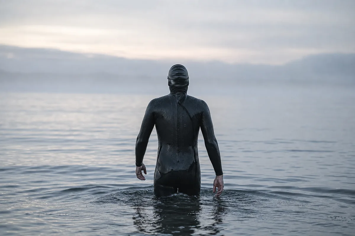 Nageur en combinaison néoprène vu de dos entrant dans une mer grise au petit matin